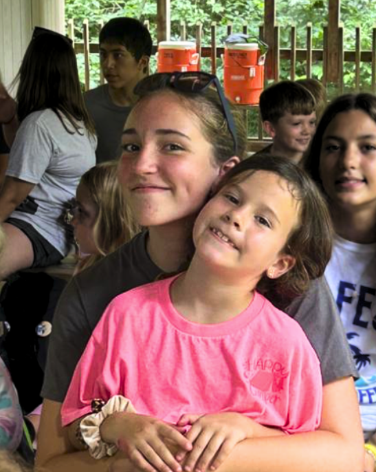 A smiling older girl hugs a younger girl in a bright pink shirt while sitting among a group of children at an outdoor gathering or camp. Trees and orange water coolers are visible in the background.