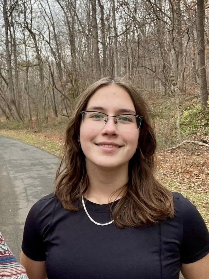 A young person with long brown hair and glasses smiles while standing on a paved path in a forested area with bare trees and fallen leaves on the ground.