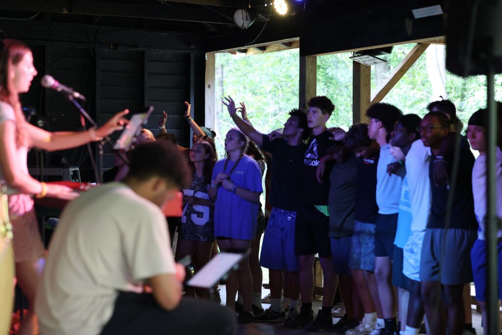 A group of teenagers stands closely together indoors at a Christian Summer Camp, some raising their hands and others watching a woman sing and play keyboard on stage. Natural light comes through an open wall behind them.