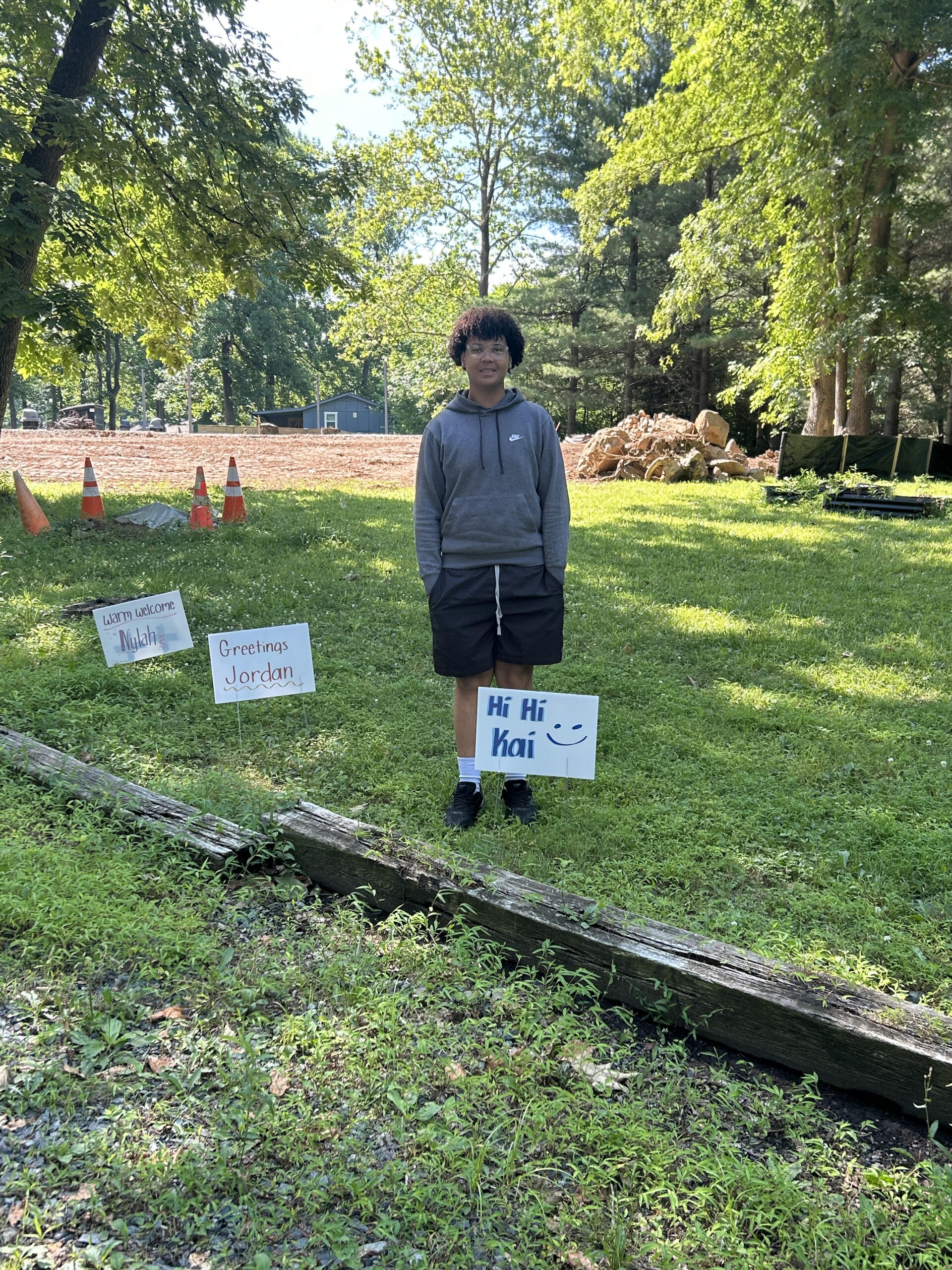 A person stands outside on grass holding a Hi Hi Kai! sign. Nearby are two lawn signs saying Greetings Jordan and Lastly, Lillith. Trees and traffic cones are visible in the background.