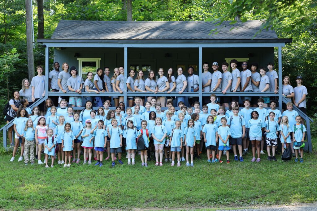A large group of children and adults in matching light blue shirts pose outside a cabin in a wooded area, smiling for a group photo on a sunny day.