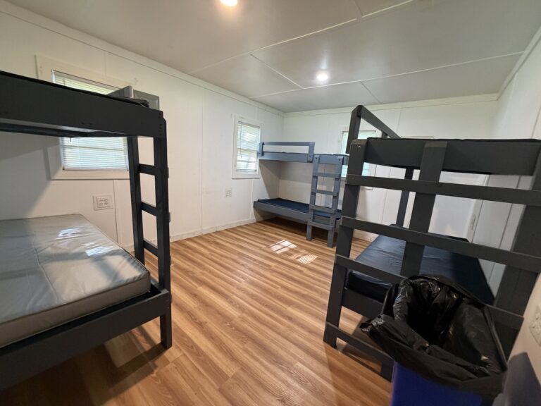 A room with three blue metal bunk beds, each with mattresses, white walls, wood-style flooring, two windows, and a black trash bin in the corner.