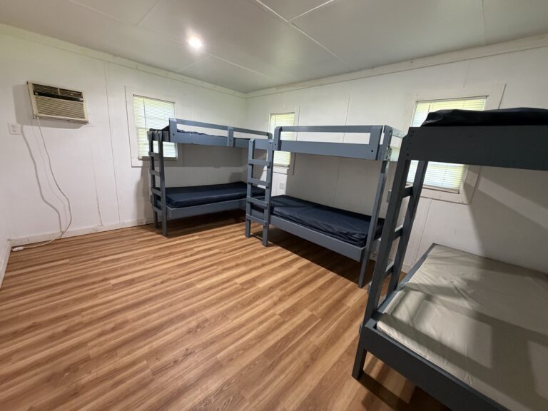 A simple room with three blue bunk beds, bare mattresses, wood-style flooring, white walls, and an air conditioner mounted on the wall.
