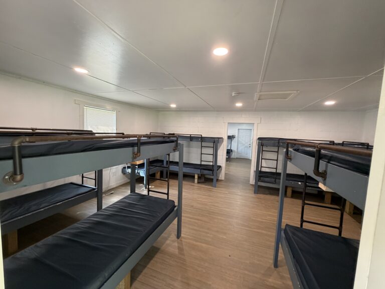 A clean room with wooden floors contains six blue metal bunk beds with dark mattresses, arranged neatly. There is a window on the left wall and a closed door at the far end of the room.