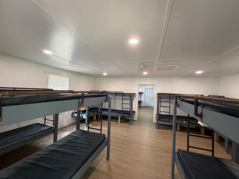 A clean dormitory room with six metal bunk beds, wood flooring, white walls, a window on the left, and a door at the far end leading to another room.
