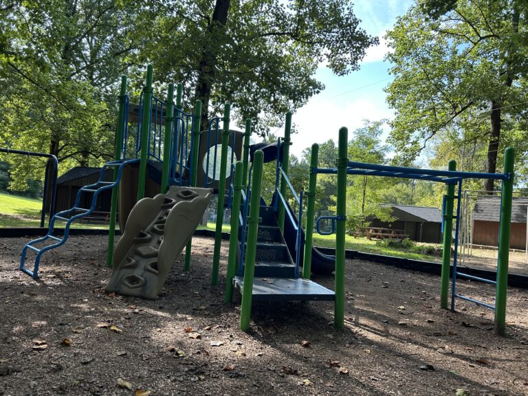 A playground with blue and green climbing structures, a slide, and a rock wall, surrounded by trees and shaded by sunlight. There are picnic tables and buildings in the background.