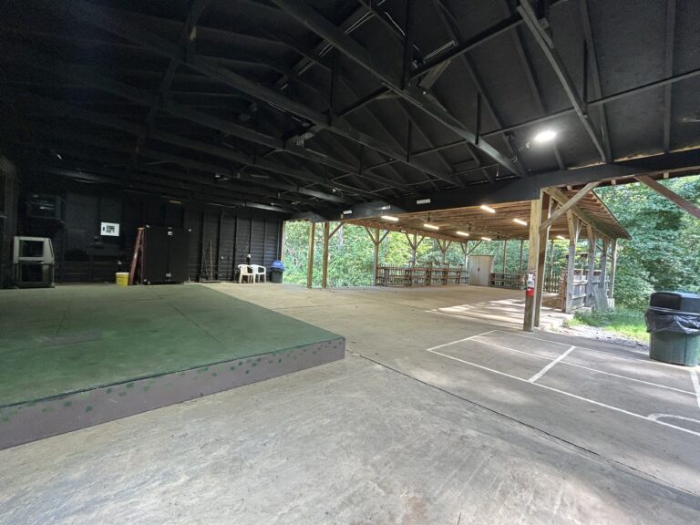 A large open-air pavilion with a small raised stage, concrete floor, wooden roof, and open sides overlooking trees; benches and a trash can are visible.