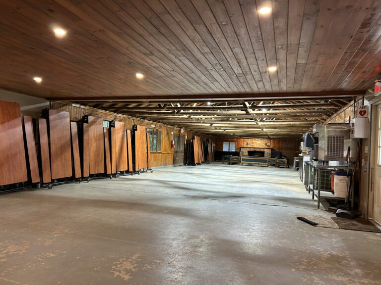 A large, empty wooden hall with folded tables stacked along the left wall, exposed beams, overhead lights, a fireplace in the back, and equipment on the right side.
