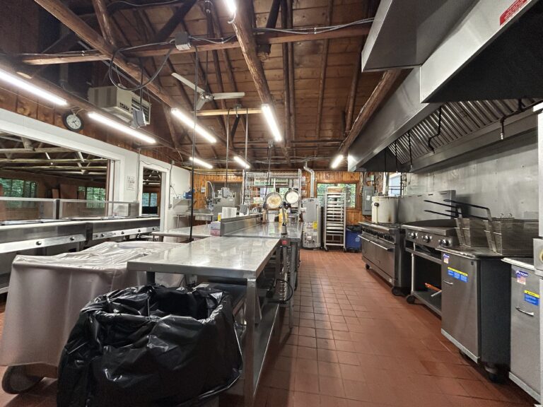 A spacious commercial kitchen with stainless steel counters, stoves, ovens, metal shelves, and a large trash bin, featuring exposed wooden beams and tile flooring.