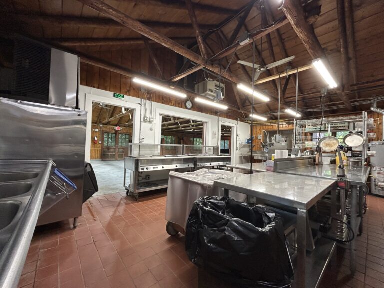 Commercial kitchen with stainless steel counters, sinks, and appliances. Exposed wooden beams and ceiling lights above. Red tile floor. Large windows and doors in the background. Black trash bin under a counter.