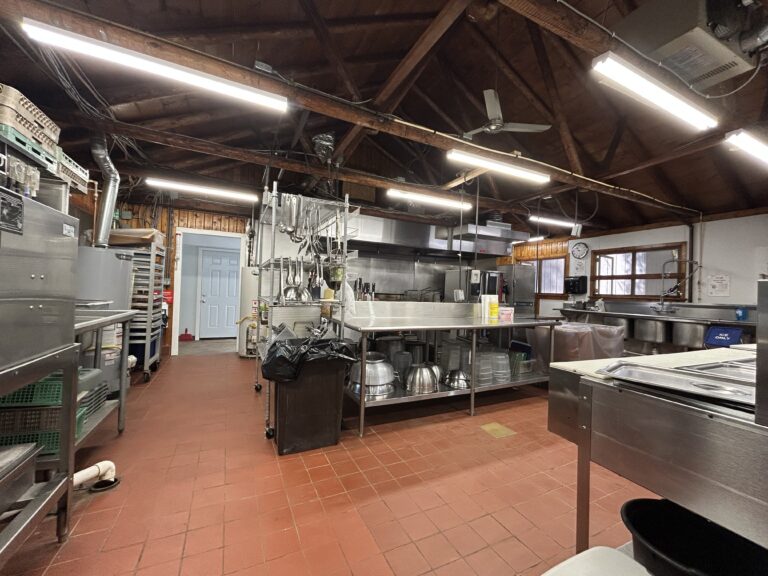 Commercial kitchen with red tile floor, stainless steel counters, shelves, sinks, and industrial equipment. Exposed beams and bright ceiling lights. Mixing bowls and racks are visible, creating an organized workspace.