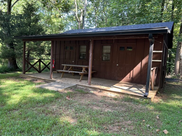 A small brown wooden cabin with a covered porch, picnic table, and fire extinguisher sits surrounded by green grass and trees on a sunny day.