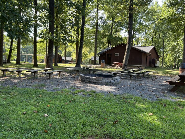 A circular fire pit surrounded by wooden benches in a forested area, with a brown cabin and picnic tables in the background on a sunny day.