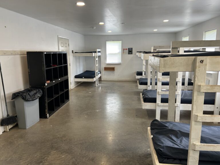 A dormitory room with six white wooden bunk beds, a black cubby storage unit, a trash can, and windows letting in natural light. The floor is concrete and the room appears clean and organized.