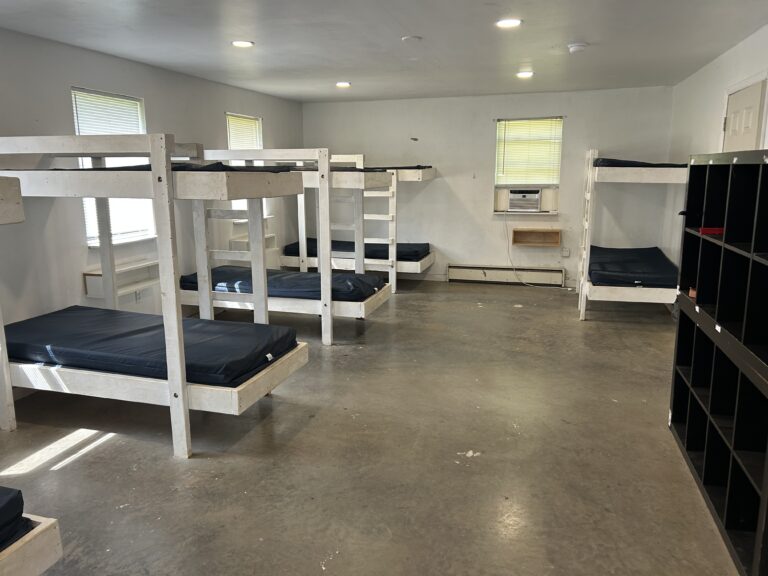 A dorm room with six white wooden bunk beds, blue mattresses, a black cubby shelf, three windows with blinds, an air conditioner, and a concrete floor.